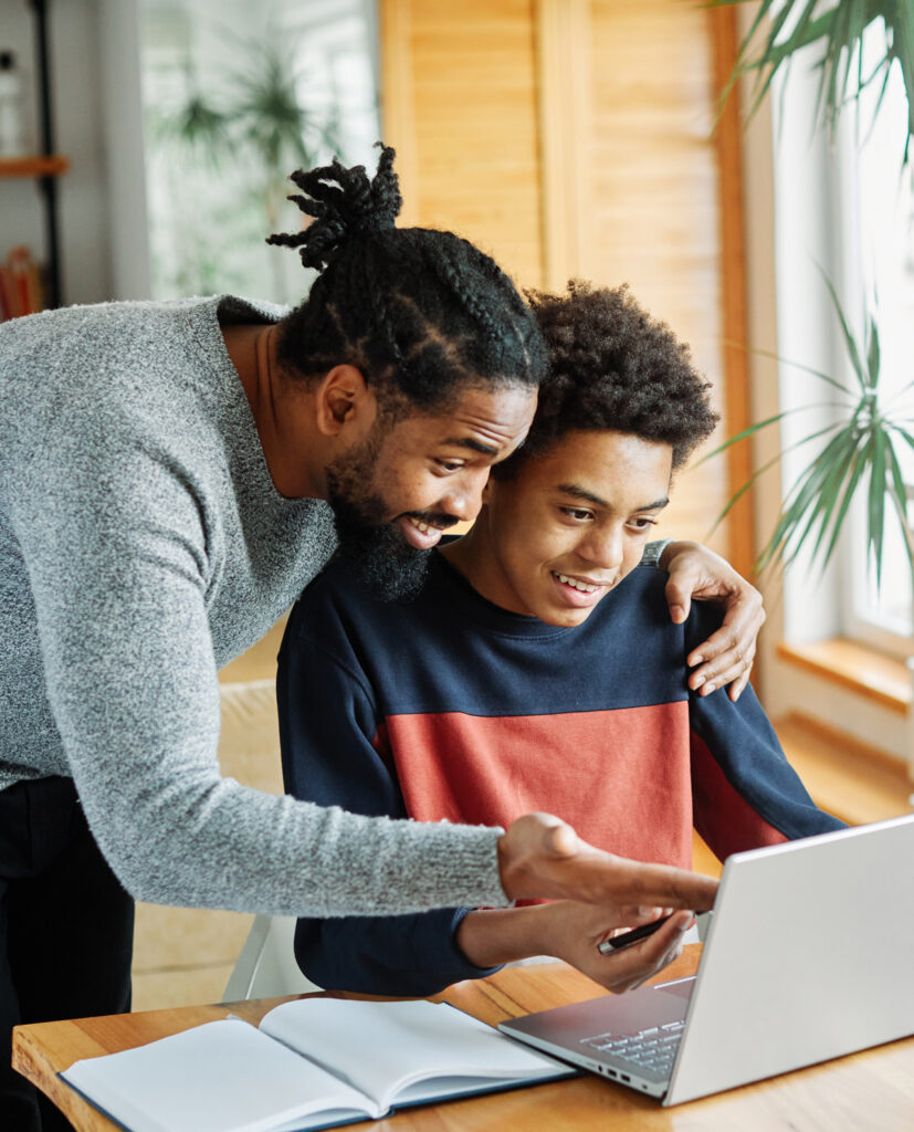 Son on laptop with his dad over his shoulder pointing at the laptop