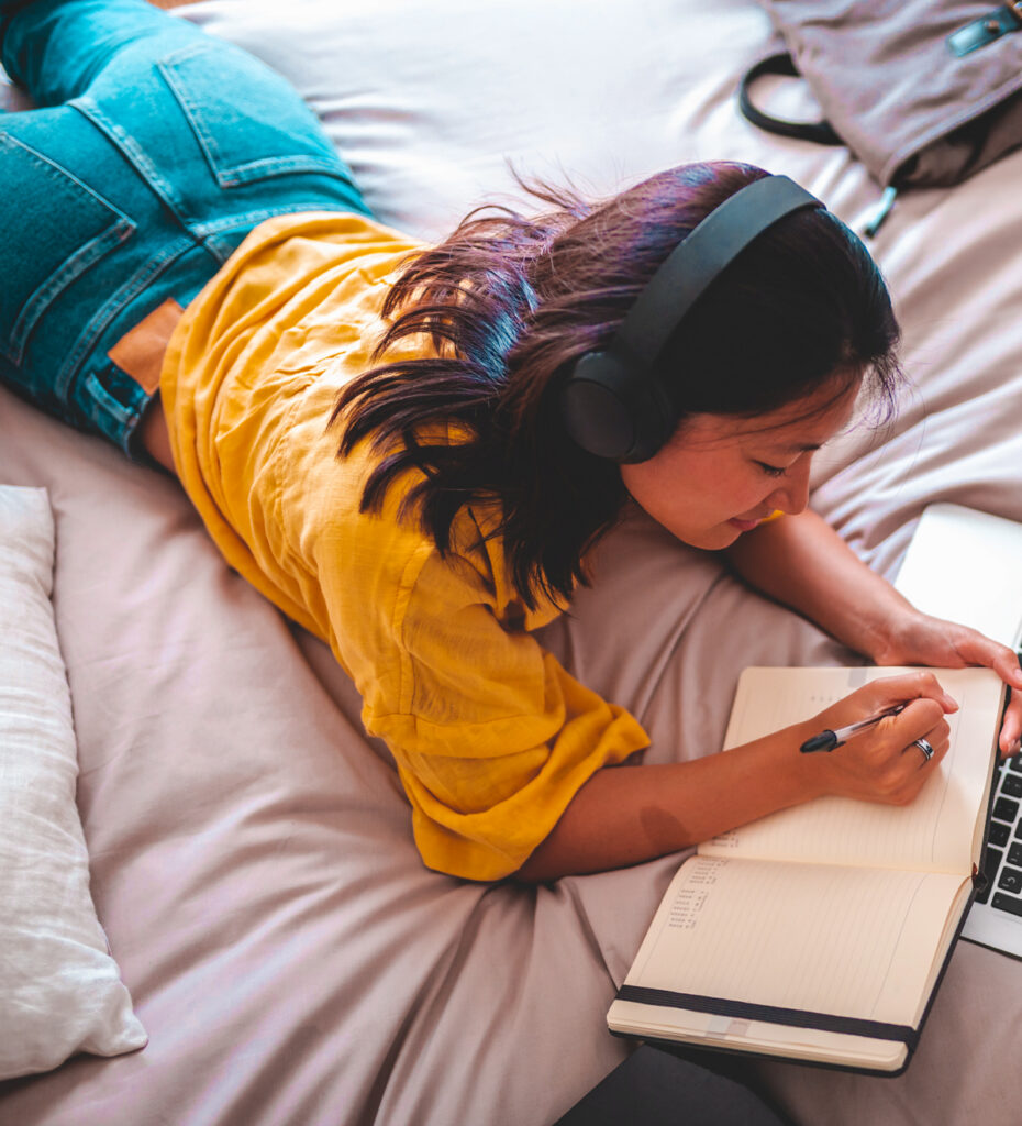 woman laying on her bed with headphones on and writing in journal