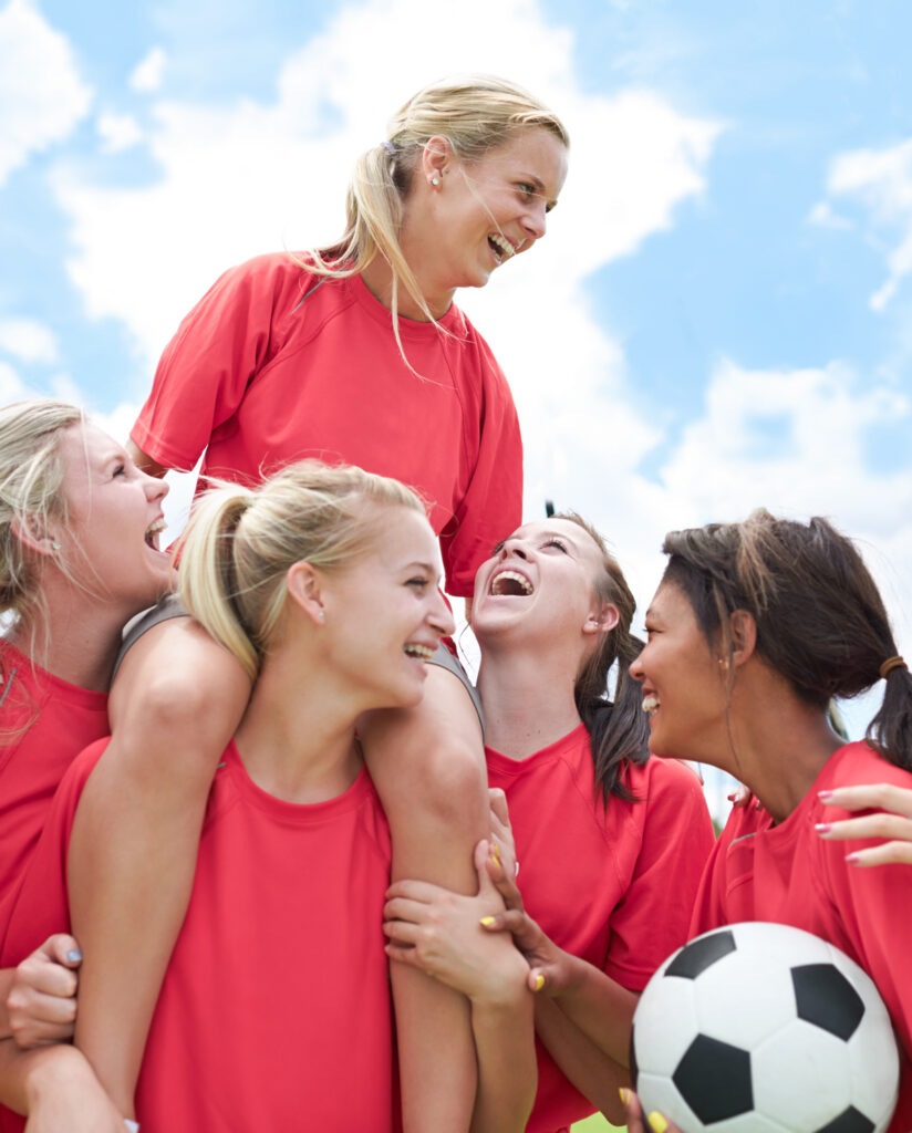 Five girls in red shirts on soccer team, one girl on another's shoulders, everyone is laughing.